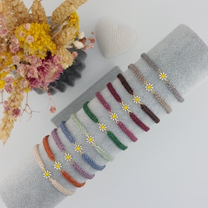Könnte beinhalten: Eine Auswahl an Armbändern mit Gänseblümchen-Anhängern in verschiedenen Farben, darunter Orange, Blau, Grün und Braun. Jedes Armband hat ein gewebtes Band und einen goldfarbenen Gänseblümchen-Anhänger. Die Armbänder werden auf einer grauen Oberfläche präsentiert, mit Trockenblumen und einer herzförmigen Dekoration im Hintergrund.
