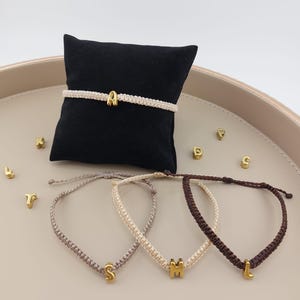 Könnte beinhalten: Eine Auswahl an personalisierten Makramee-Armbändern mit goldfarbenen Initialen-Anhängern. Ein Armband wird auf einem schwarzen Kissen präsentiert, während drei weitere darunter angeordnet sind. Zusätzliche goldene Buchstaben sind verstreut.