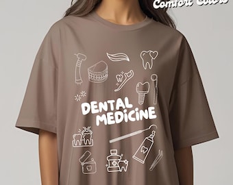 Op de afbeelding: Een taupekleurig T-shirt met witte lijntekeningen van tandheelkundige instrumenten en tanden. De woorden "DENTAL MEDICINE" zijn in witte blokletters gedrukt. De rechterbovenhoek van het shirt heeft de woorden "Pepper Comfort Colors" in een cursief lettertype.