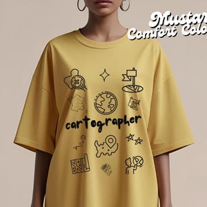 May include: Mustard-colored t-shirt with the word "cartographer" and various map-related illustrations in black. The shirt also has the text "Mustard Comfort Colors" in a script font.