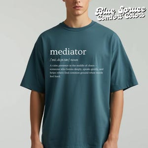 May include: Teal-colored t-shirt with the word "mediator" printed in white, along with its phonetic pronunciation and definition. The text "Blue Spruce Comfort Colors" is in the upper right corner. The shirt is a short-sleeved, crew neck style.