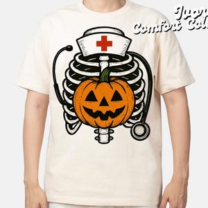 May include: Cream-coloured t-shirt with a Halloween design. The graphic shows a skeleton torso, a jack-o'-lantern, a nurse's hat with a red cross, and a stethoscope. The words "Juicy Comfort Colors" are printed on the upper right.
