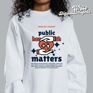 以下が含まれることがあります： 白いスウェットシャツに、「Speak Out, Stand Up Public Health Matters」というグラフィックデザインが施されています。グラフィックには、プラス記号と人が入ったハートが描かれており、星で囲まれています。