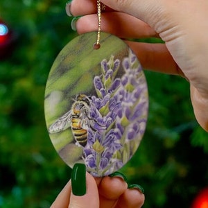 Może przedstawiać: Owalna ozdoba ceramiczna z pszczołą na fioletowych kwiatach. Ozdoba ma złotą sznurkę do zawieszenia.