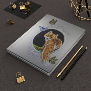 May include: A silver journal with a fox illustration in a circular frame. The fox is looking to the left and has a bushy tail. There are butterflies on the cover, and the journal has a Celtic knot design.