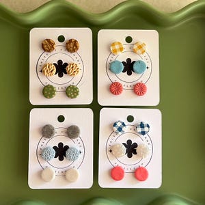 Peut inclure: Quatre ensembles de boucles d'oreilles clous faites à la main sur des cartes blanches. Chaque carte présente trois paires de boucles d'oreilles de différentes couleurs et motifs, dont du vichy, floral et à pois. Les boucles d'oreilles sont présentées sur un plateau vert.