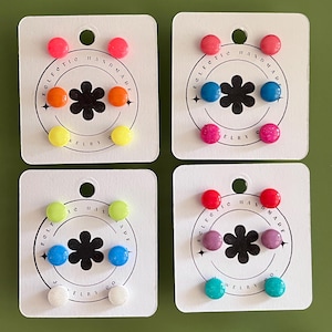 Op de afbeelding: Vier sets kleurrijke oorstekers op witte kaarten. Elke kaart heeft drie paar oorbellen in verschillende kleuren, waaronder roze, oranje, geel, blauw, paars en groen. De kaarten hebben een bloemenontwerp en de tekst "Felfe Handmade".