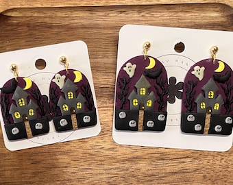 May include: Two pairs of arch-shaped dangle earrings featuring a haunted house scene with a purple sky, yellow crescent moon, white ghost, black bat, and bare trees. The base is black with small gray tombstones marked with the letters RIP.