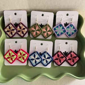 Op de afbeelding: Een collectie handgemaakte polymeerklei oorbellen in verschillende kleuren, waaronder roze, groen, blauw en paars. Elk paar heeft een geometrisch, op quilts geïnspireerd ontwerp met goudkleurige oorhaken. De oorbellen worden op witte kaarten gepresenteerd.