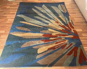 May include: A rectangular rug with a floral design. The rug has a dark blue background and a pattern of stylized, radiating shapes in shades of brown, tan, red, and grey. It is placed on a wooden floor.