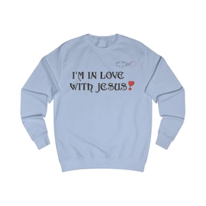 Könnte beinhalten: Hellblaues Sweatshirt mit dem Text "I'M IN LOVE WITH JESUS" in schwarzer Schrift. Ein rotes Herz-Symbol befindet sich neben dem Wort "JESUS". Oberhalb des Textes befindet sich eine kleine Grafik mit den Worten "LOVE LOVES LOVE".