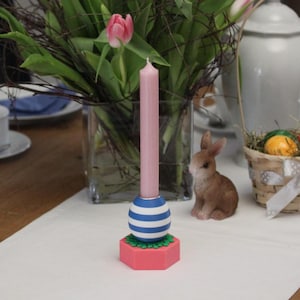 Może przedstawiać: Różowa świeca w niebiesko-białym, pasiastym świeczniku, ustawiona na różowej, sześciokątnej podstawie z zielonymi obramowaniami. Mała brązowa figurka królika i kosz z jajkiem w tle. Widoczny jest również wazon z tulipanami.