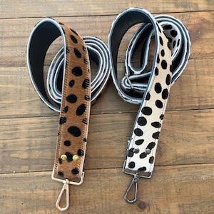 May include: Two adjustable bag straps with a textured animal print pattern. One strap features a tan base with black spots and gold-toned hardware, while the other has a white base with black spots and dark gunmetal-toned hardware.
