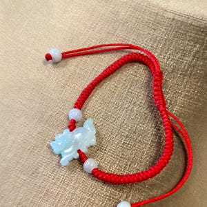 May include: A red string bracelet with a jade green pendant in the shape of a dragon. The bracelet has white beads and is tied with a knot.