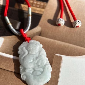 May include: A jade pendant necklace with a carved jade figure of a mythical creature. The pendant is attached to a red cord with white beads. The necklace is displayed in a white box.