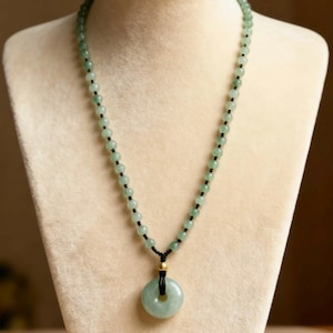 May include: A necklace featuring light green, round, polished stone beads strung on a black cord. The necklace includes a central circular green stone pendant, known as a donut, suspended by a black cord with a small, shiny gold-toned bead.