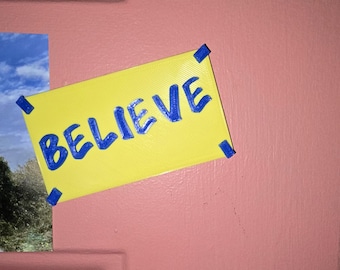 May include: A yellow rectangular sign with the word "BELIEVE" in blue lettering is taped to a pink door. The sign is secured with blue tape in each corner. A partial image of a landscape is visible to the left.