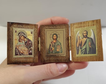 Könnte beinhalten: Ein kleines, aufklappbares Holzikon mit drei Tafeln, die religiöse Bilder zeigen. Die Tafeln zeigen Figuren mit goldenen Akzenten, gerahmt in dunklem, verziertem Holz. Das Ikon ist geöffnet und zeigt die detaillierten Kunstwerke.
