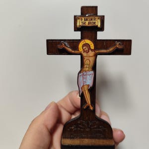 Puede incluir: Un crucifijo de madera marrón oscuro con una imagen pintada de Jesucristo. La cruz tiene una base rectangular. La imagen de Jesús está en el centro, con el texto "O BACIΛEYC THC AOΞHC" arriba.
