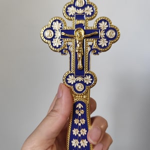 May include: An ornate Orthodox cross in gold and blue, featuring a depiction of Jesus Christ. The cross has detailed floral patterns and religious text. The cross is held in a hand.