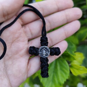 May include: A black woven cross pendant necklace with a silver-coloured medallion at the centre. The cross is made of knotted cord, and the medallion has a detailed design. The necklace is shown against a blurred green background.