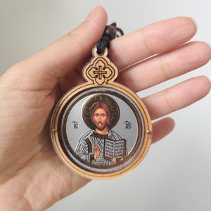 May include: Circular wooden pendant with an icon of Jesus Christ. The icon is set within a circular frame, with a detailed image of Jesus holding a book. The pendant has a decorative wooden top with a cross design and a black cord.