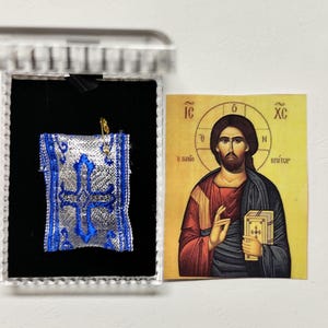 Może przedstawiać: Niebieski i srebrny haftowany przedmiot religijny w przezroczystym pudełku, obok ikony religijnej przedstawiającej mężczyznę z aureolą, ubranego w czerwone i niebieskie szaty, trzymającego książkę. Ikona ma grecki tekst.