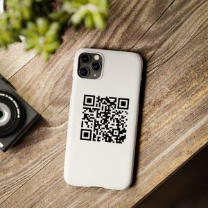 May include: White iPhone case with a black QR code printed on the back. The case has a camera lens cutout in the top left corner.