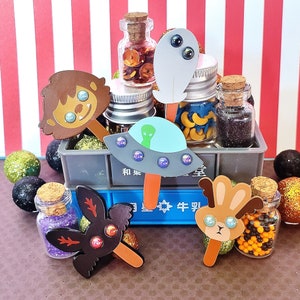 May include: A collection of colorful, whimsical crafts, including a UFO, a monster, a bunny, and a cat, all made with paper and embellishments. The crafts are displayed on a wooden shelf with a striped background.