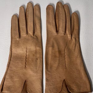 K&ouml;nnte beinhalten: Ein Paar hellbraune Lederhandschuhe. Die Handschuhe haben ein klassisches Design mit einzelnen Fingerschlitzen und gen&auml;hten N&auml;hten an den Seiten. Das Leder hat ein leicht strukturiertes Aussehen und einen warmen, neutralen Farbton.