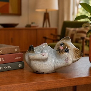May include: A ceramic cat figurine in a relaxed pose, lying on its back. The cat has a speckled gray body, brown head, and pink paw pads. The figurine is on a wooden surface, with books and a plant in the background.
