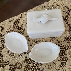 Pode incluir: Uma caixa de cerâmica branca com um design floral decorativo na tampa. A caixa está sobre uma toalha de renda branca. Há dois pequenos pratos de cerâmica branca ao lado da caixa.