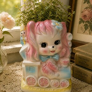 May include: A vintage-style ceramic planter shaped like a puppy with pink hair and a blue bow. The planter is decorated with a train design and filled with green plants. The color palette includes pastel pink, blue, and yellow.