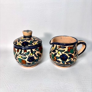 May include: A ceramic sugar bowl with a lid and a creamer. Both pieces are decorated with a floral pattern in blue, red, orange, and green on a cream background. The rims and handles are dark blue.