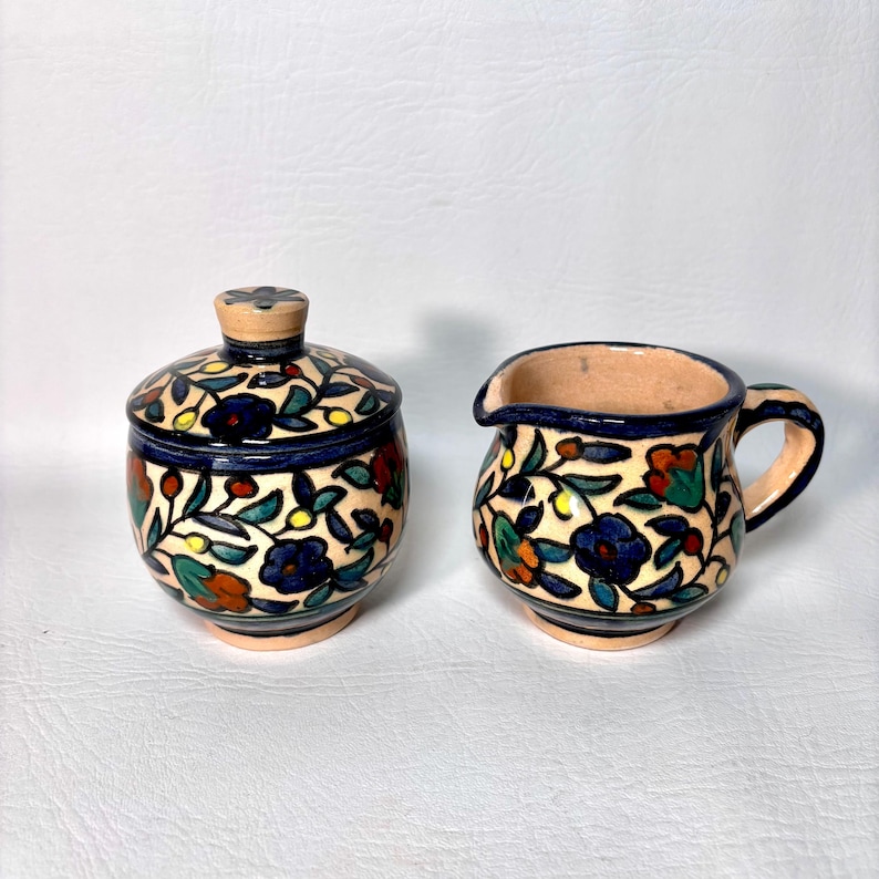 May include: A ceramic sugar bowl with a lid and a creamer. Both pieces are decorated with a floral pattern in blue, red, orange, and green on a cream background. The rims and handles are dark blue.