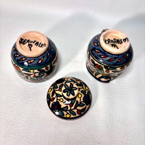 May include: Three ceramic pieces with hand-painted floral designs. Two small cups and a lid are visible. The cups have blue, green, and red accents, with the word "Jerusalem" written on the bottom. The lid features a floral pattern.