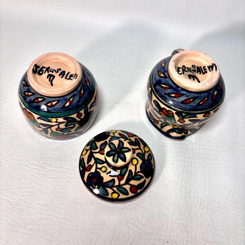 May include: Three ceramic pieces with hand-painted floral designs. Two small cups and a lid are visible. The cups have blue, green, and red accents, with the word "Jerusalem" written on the bottom. The lid features a floral pattern.