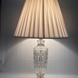 May include: A clear crystal table lamp with a pleated white lampshade. The lamp base is intricately cut with a diamond pattern.