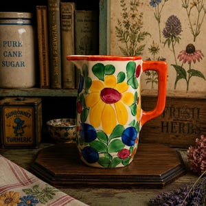 May include: A hand-painted ceramic pitcher with a vibrant floral design. The pitcher features a large yellow sunflower with a red center, blue berries, and green leaves. It has an orange handle and rim, and sits on a wooden surface.