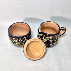 May include: A ceramic creamer and sugar bowl set with a floral design. The set includes a small pitcher with a handle, a round bowl, and a lid. The pieces are beige with blue, red, and green floral patterns and a dark blue trim.