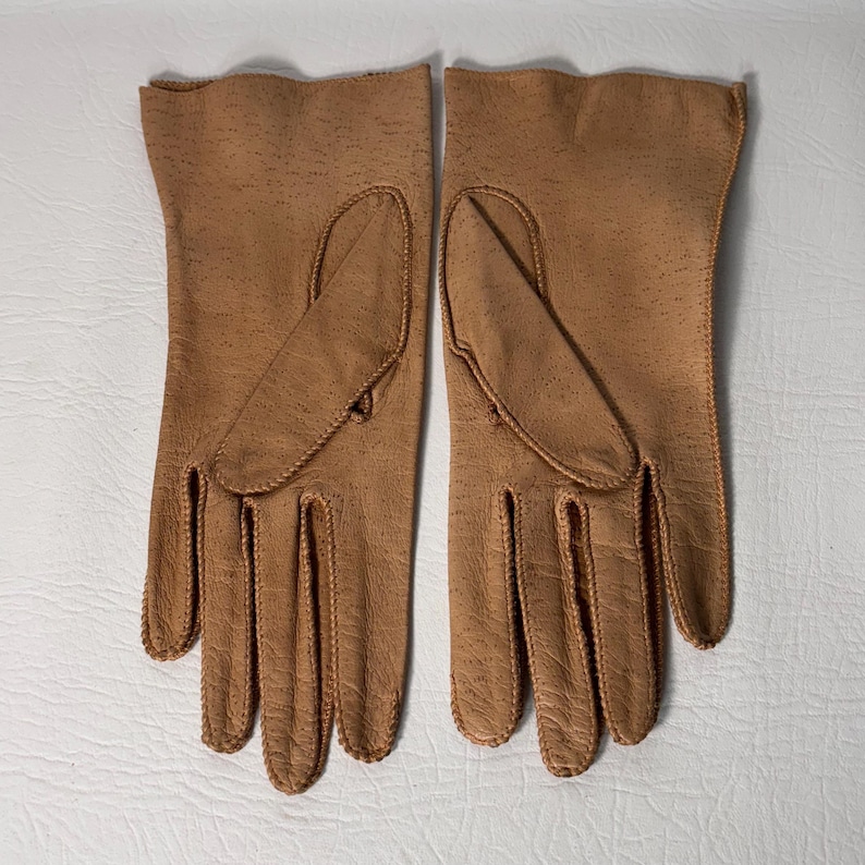 K&ouml;nnte beinhalten: Ein Paar hellbraune Lederhandschuhe, flach ausgelegt. Die Handschuhe haben eine strukturierte Oberfl&auml;che und sind mit passendem Faden gen&auml;ht. Die Handschuhe haben ein klassisches Design mit separatem Daumen und Fingern.