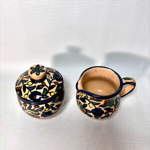 May include: A ceramic sugar bowl with a lid and a creamer. Both pieces are decorated with a floral pattern in blue, green, red, and yellow on a cream background. The sugar bowl has a round shape, and the creamer has a handle and a spout.
