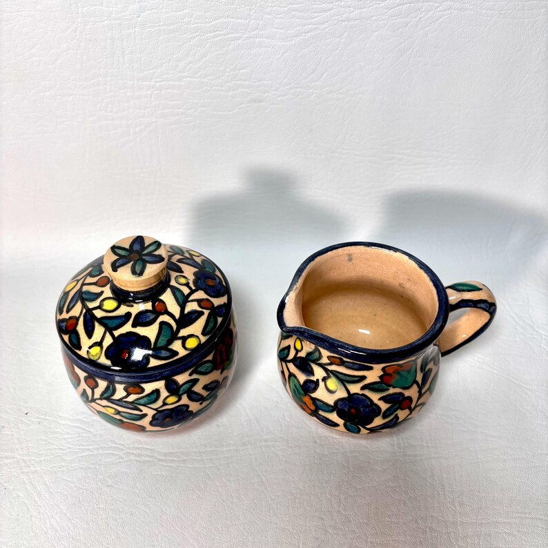 May include: A ceramic sugar bowl with a lid and a creamer. Both pieces are decorated with a floral pattern in blue, green, red, and yellow on a cream background. The sugar bowl has a round shape, and the creamer has a handle and a spout.