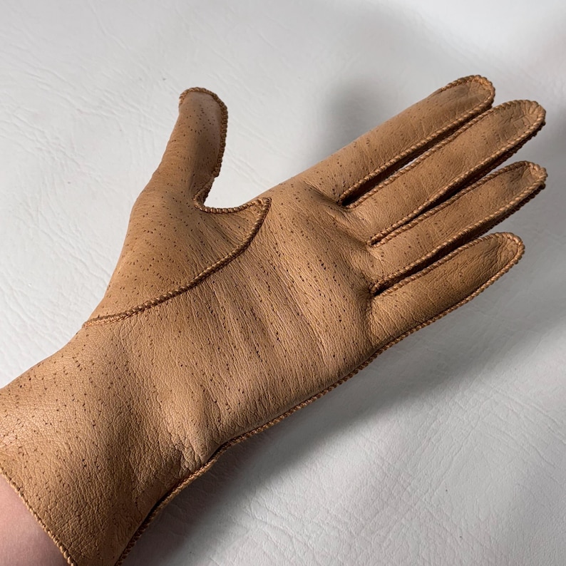 K&ouml;nnte beinhalten: Hellbrauner Lederhandschuh mit strukturierter Oberfl&auml;che und dekorativen N&auml;hten. Der Handschuh ist im klassischen F&uuml;nf-Finger-Stil gehalten und zeigt eine Vintage-&Auml;sthetik.