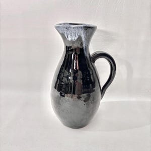 Puede incluir: Una jarra de cerámica negra con acabado brillante. La jarra tiene un asa y un borde acampanado con un esmalte gris y blanco. El cuerpo de la jarra refleja el entorno. Una pieza decorativa para el hogar.
