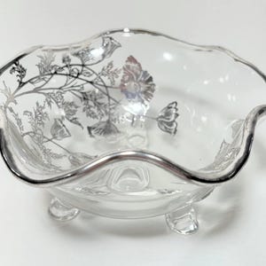 May include: A clear glass bowl with a silver floral design and three legs. The bowl has a scalloped edge.