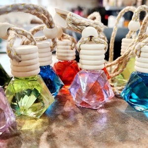 May include: A collection of six colorful, diamond-shaped glass bottles with wooden stoppers and twine hangers. The bottles are filled with different colored liquids and are arranged in a row on a wooden surface.