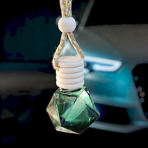 May include: A green glass car air freshener in a geometric shape hangs from a beige rope with wooden beads. The air freshener is hanging in front of a blue car.