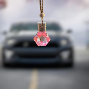May include: A pink diamond-shaped car air freshener hanging from a string with a wooden bead. The air freshener is in focus, while a car is blurred in the background.
