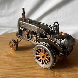 May include: A detailed metal tractor model with silver and copper accents. The miniature tractor features a steering wheel, seat, and spoke wheels. The model is displayed on a wooden surface.